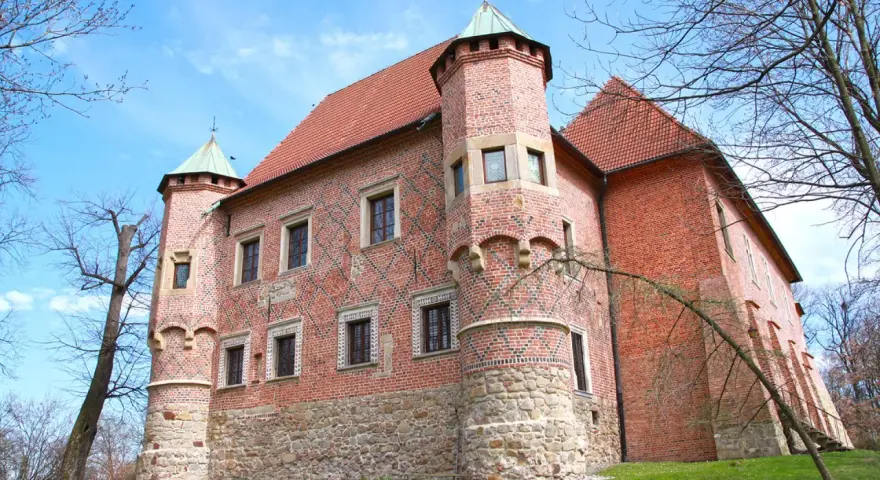 Zamek w Dębnie, ceglana twierdza z kamienną podstawą i zielonymi wieżyczkami, otoczony drzewami pod błękitnym niebem.