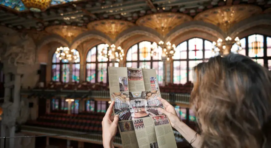 Kobieta studiuje broszurę z widokiem na wspaniałe wnętrze Pałacu Muzyki Katalońskiej, z witrażowym sufitem i rzędami siedzeń.