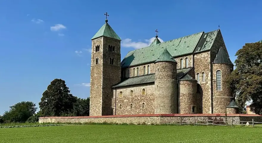 Kamienny zamek w Tumie z zielonym dachem, otoczony murem i zieloną łąką, pod błękitnym niebem.
