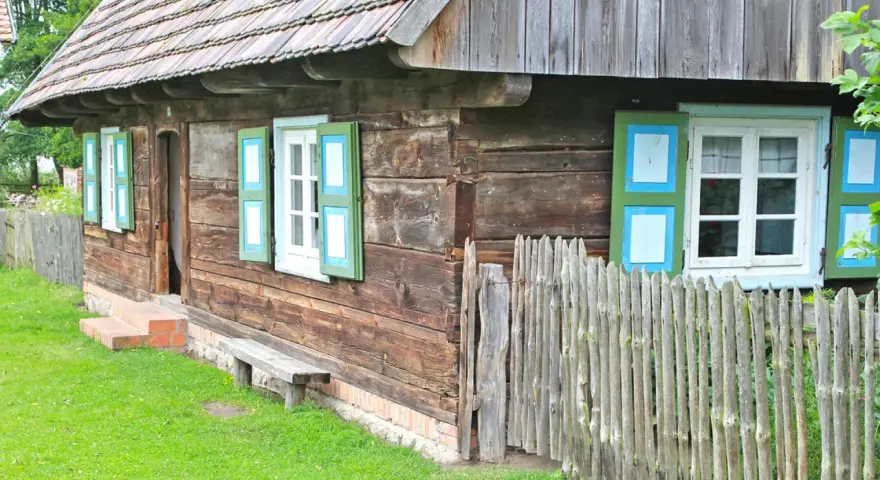 Drewniana chata z zielono-niebieskimi okiennicami, ławką i płotem. Skansen w Dziekanowicach zaprasza do podróży w czasie.