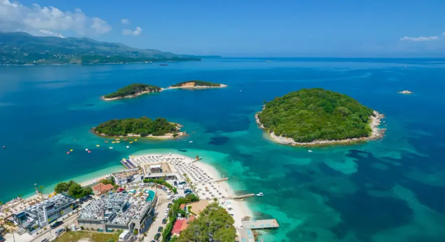 Albania Durrës atrakcje: malownicze wysepki z zieloną roślinnością otoczone turkusową wodą, plaża z leżakami i budynkami hotelowymi.