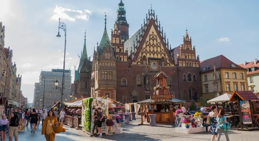 Tłum ludzi spaceruje po starym rynku we Wrocławiu, podziwiając piękną architekturę i letnie stoiska.