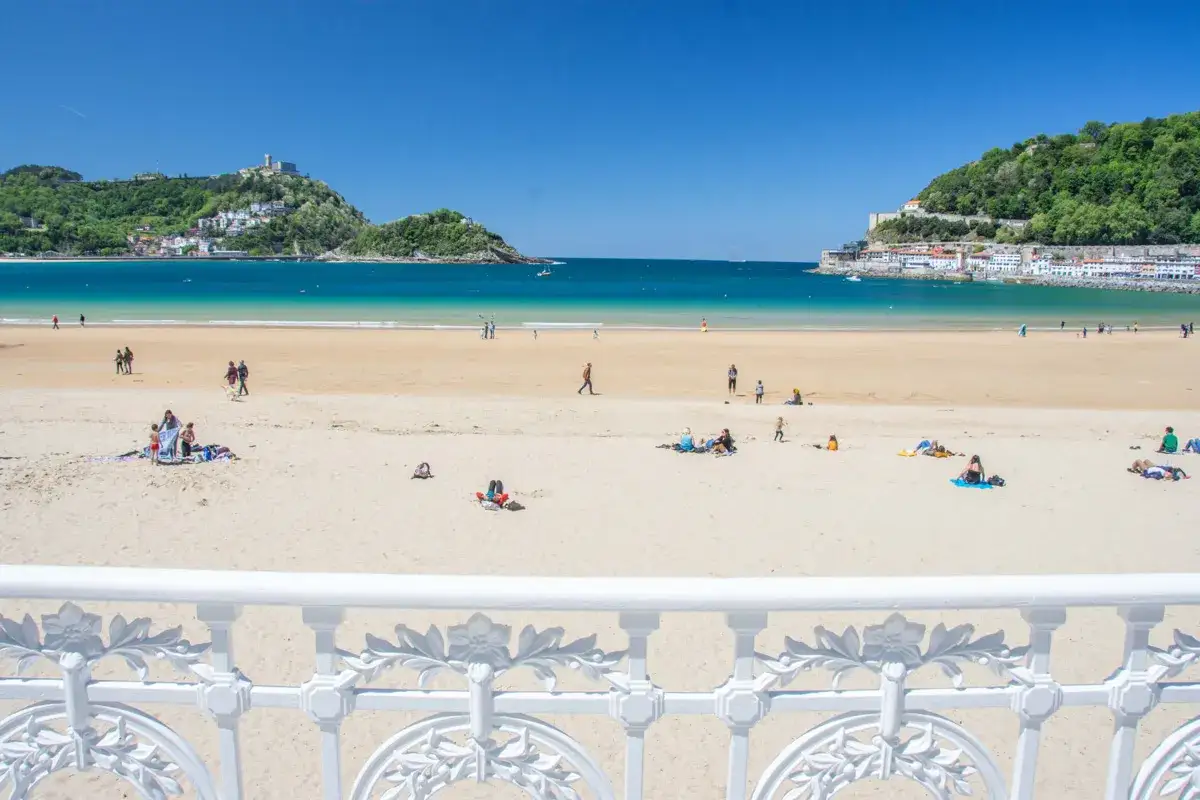 Słoneczna plaża w San Sebastian z turkusową wodą i zielonymi wzgórzami w tle. Ludzie odpoczywają na piasku, ciesząc się wakacjami.