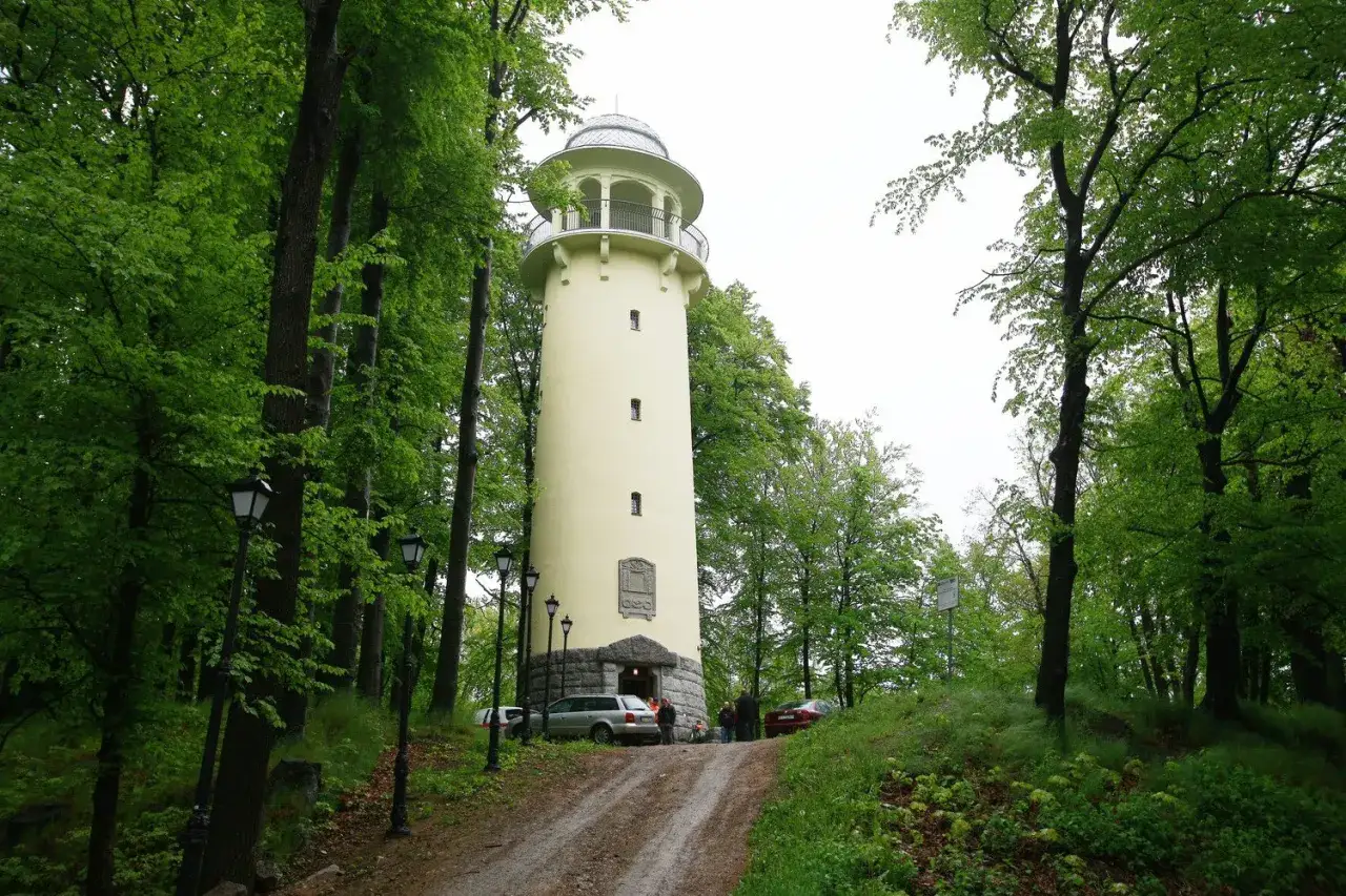 Jasnoż&oacute;łta wieża widokowa w Jeleniej G&oacute;rze, otoczona bujną zielenią drzew. U podn&oacute;ża wieży stoją samochody i kilka os&oacute;b.