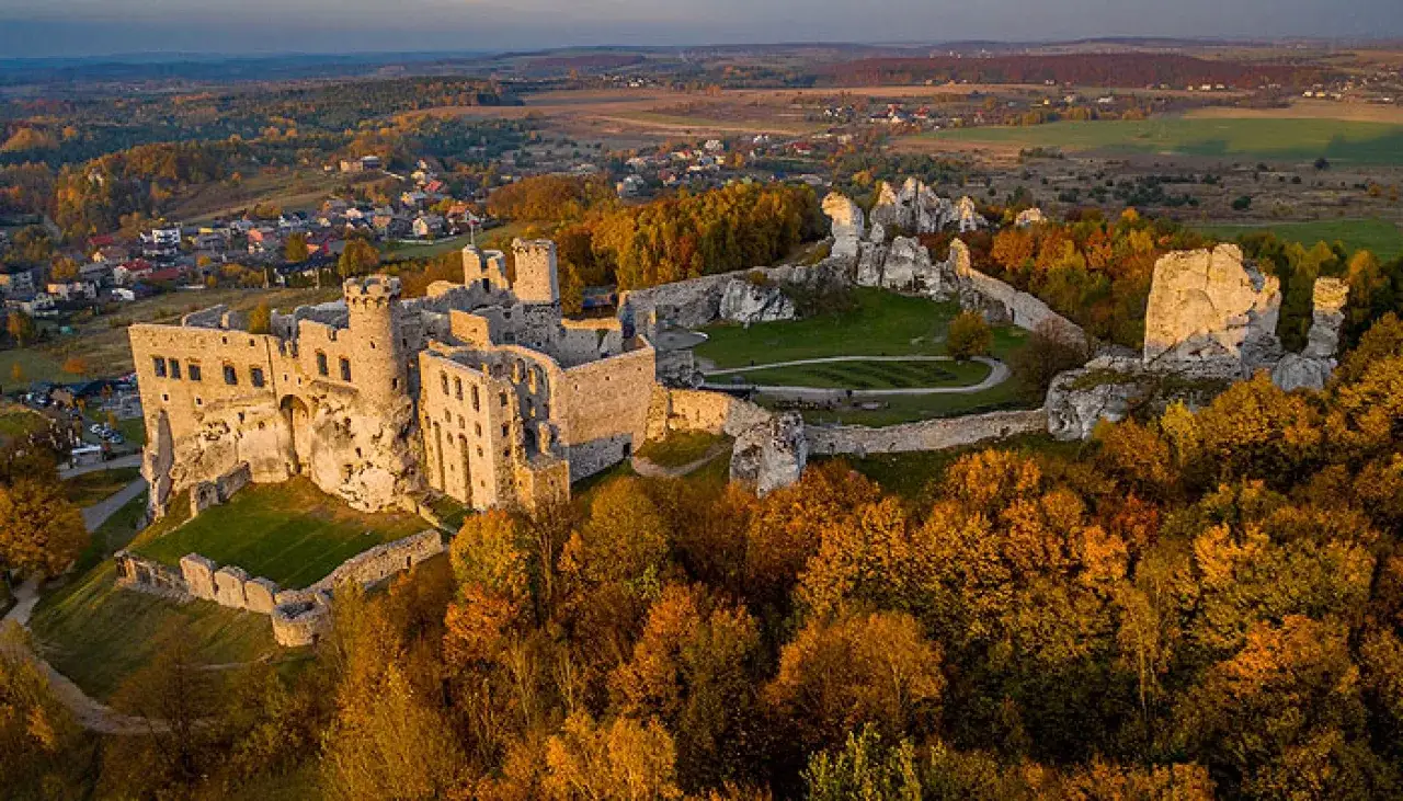 Zamek Ogrodzieniec w jesiennej szacie, otoczony kolorowymi drzewami i skalistymi ostańcami, majestatycznie g&oacute;ruje nad okolicą.