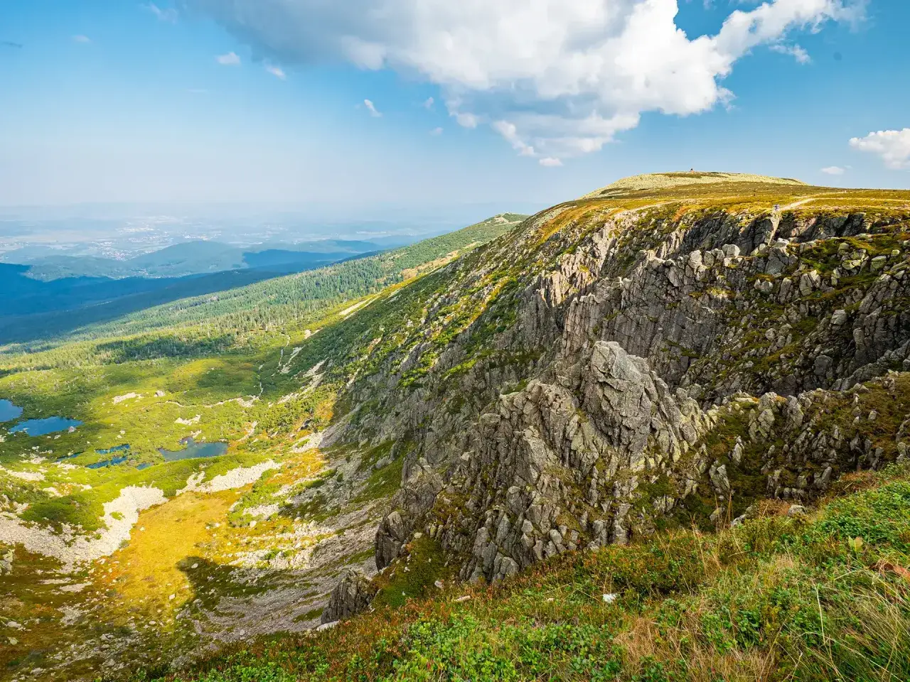 Widok na skaliste zbocze i dolinę z jeziorami, w oddali widać miasto. W tle wznosi się szczyt, gdzie znajduje się schronisko w Śnieżnych Kotłach.