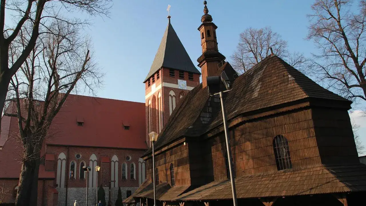 Drewniana i ceglana architektura kościoł&oacute;w w miasteczku śląskim, z wieżą zegarową i strzelistymi dachami, pod błękitnym niebem.