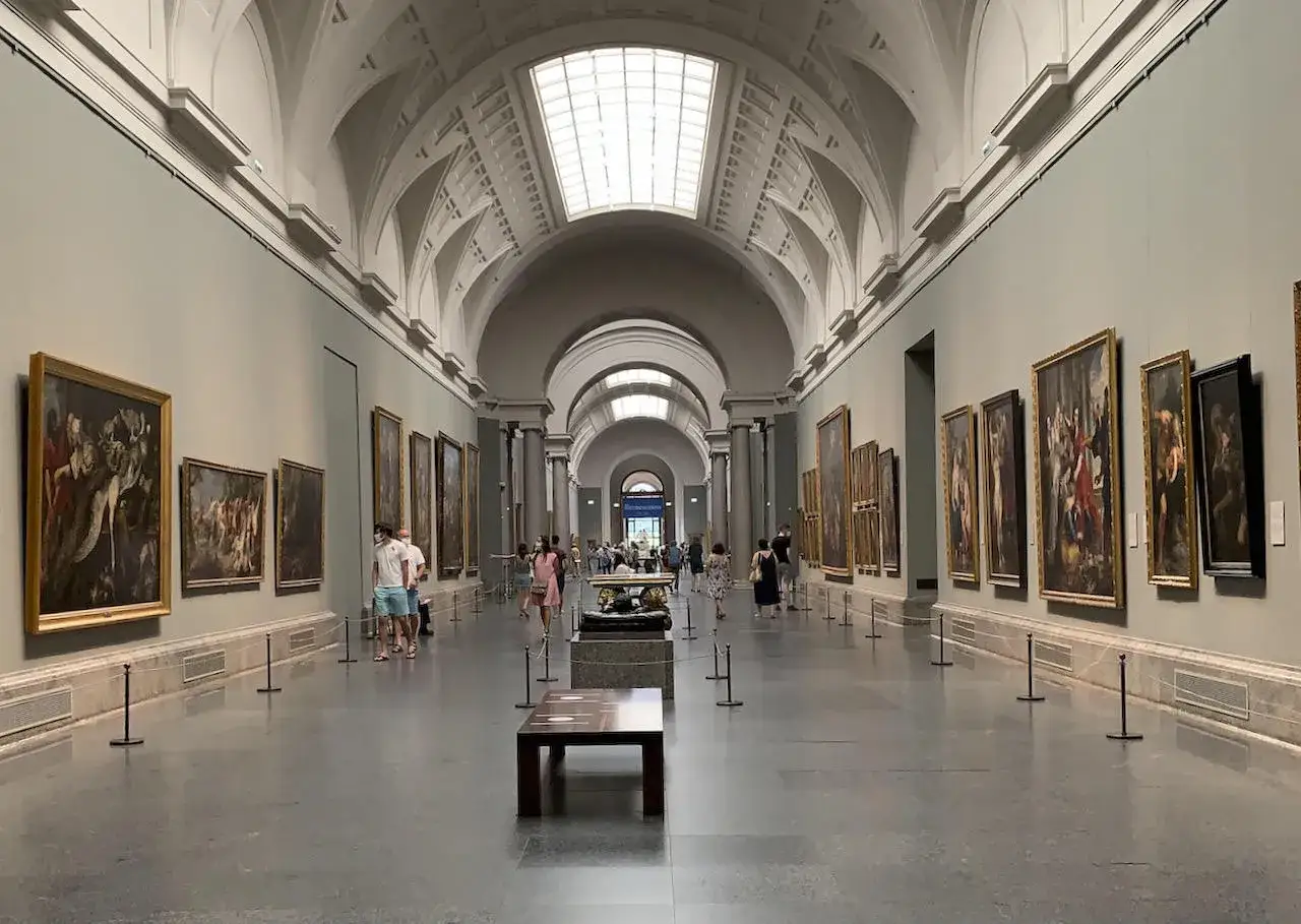 Długa, jasna sala pełna obraz&oacute;w w złotych ramach. Ludzie spacerują po muzeum w Madrycie, podziwiając dzieła sztuki.