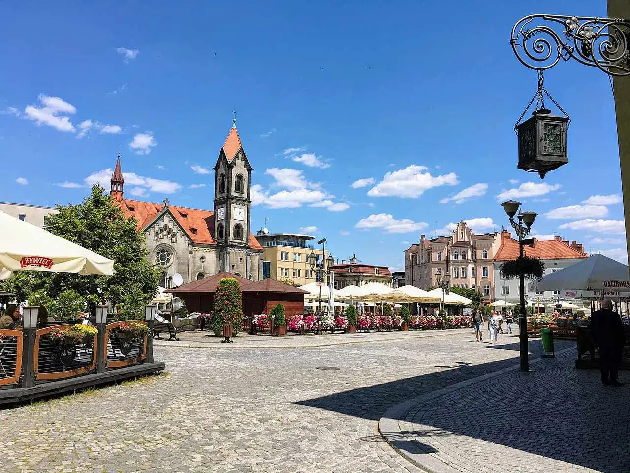 Rynek w Tarnowskich Górach tętni życiem. Widać kościół, kawiarniane ogródki i ludzi spacerujących po bruku.
