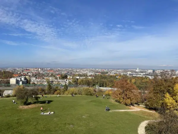 Jesienny widok na Kraków z Kopca Kraka. Ludzie odpoczywają na trawie, podziwiając panoramę miasta i kolorowe drzewa.