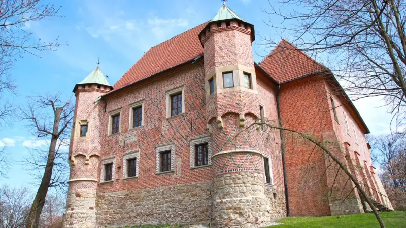 Zamek w Dębnie, ceglana twierdza z kamienną podstawą i zielonymi wieżyczkami, otoczony drzewami pod błękitnym niebem.