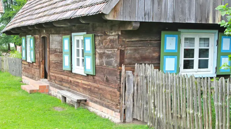Drewniana chata z zielono-niebieskimi okiennicami, ławką i płotem. Skansen w Dziekanowicach zaprasza do podróży w czasie.